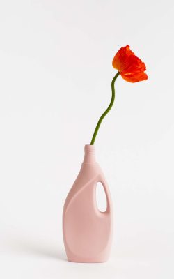 Prikazana je vaza brenda foekje fleur u svijetlo ružićastoj boji ispred sive pozadine. Vaza ima oblik kontejnera za sredstvo za pranje suđa. U vazu je utaknut crveni cvjet.