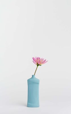 Prikazana je vaza brenda foekje fleur u svijetlo plavoj boji ispred sive pozadine. Vaza ima oblik kontejnera za sredstvo za pranje suđa. U vazu je utaknut ružićasti cvjet.