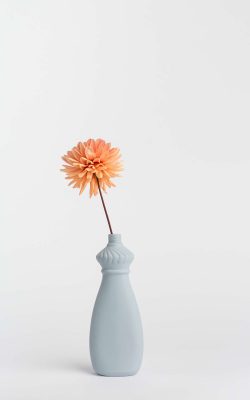 Prikazana je vaza brenda foekje fleur u svijetlo ljubičastoj boji ispred sive pozadine. Vaza ima oblik kontejnera za sredstvo za pranje suđa. U vazu je utaknut narančasti cvjet.