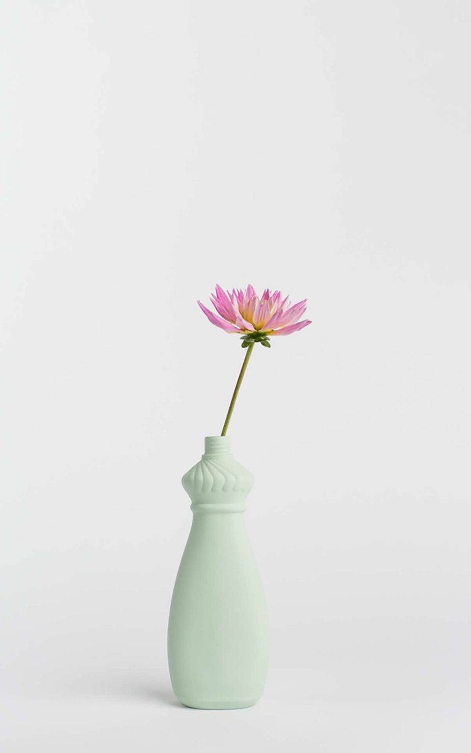 Prikazana je vaza brenda foekje fleur u svijetlo zelenoj boji ispred sive pozadine. Vaza ima oblik kontejnera za sredstvo za pranje suđa. U vazu je utaknut ružićasti cvjet.