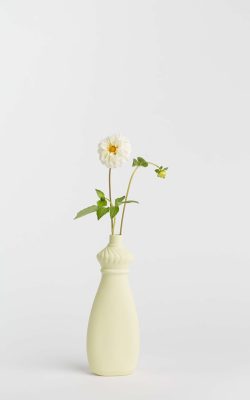 Prikazana je vaza brenda foekje fleur u svijetlo žutoj boji ispred sive pozadine. Vaza ima oblik kontejnera za sredstvo za pranje suđa. U vazu je utaknut bijeli cvjet.