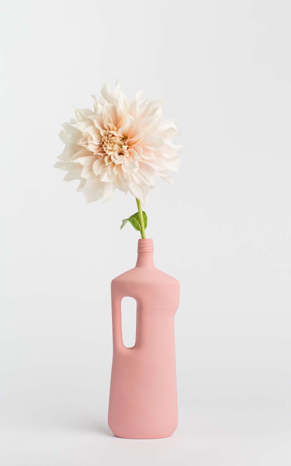 Prikazana je vaza brenda foekje fleur u svijetlo ružićastoj boji ispred sive pozadine. Vaza ima oblik kontejnera za sredstvo za pranje suđa. U vazu je utaknut bijeli cvjet.