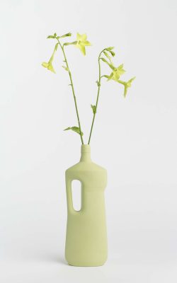 Prikazana je vaza brenda foekje fleur u svijetlo zelenoj boji ispred sive pozadine. Vaza ima oblik kontejnera za sredstvo za pranje suđa. U vazu je utaknut zeleni cvjet.