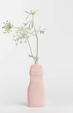 Prikazana je vaza brenda foekje fleur u svijetlo ružićastoj boji ispred sive pozadine. Vaza ima oblik kontejnera za sredstvo za pranje suđa. U vazu je utaknut zeleni cvjetić.