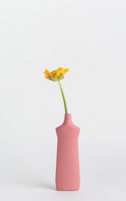 Prikazana je vaza brenda foekje fleur u ružićastoj boji ispred sive pozadine. Vaza ima oblik kontejnera za sredstvo za pranje suđa. U vazu je utaknut žuti cvjet.