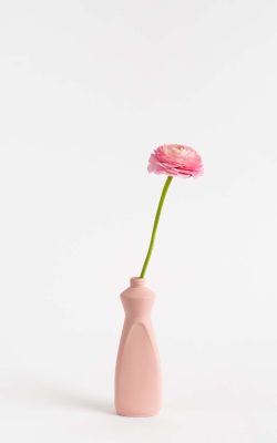 Prikazana je vaza brenda foekje fleur u svijetlo ružićastoj boji ispred sive pozadine. Vaza ima oblik kontejnera za sredstvo za pranje suđa. U vazu je utaknut ružićasti cvjet.