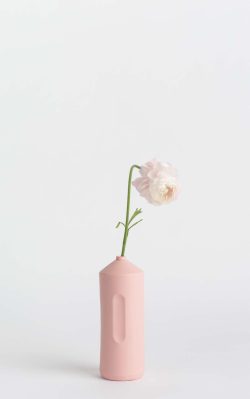 Prikazana je vaza brenda foekje fleur u svijetlo ružićastoj boji ispred sive pozadine. Vaza ima oblik kontejnera za sredstvo za pranje suđa. U vazu je utaknut bijeli cvjet.