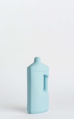 Prikazana je vaza brenda foekje fleur u svijetlo plavoj boji ispred sive pozadine. Vaza ima oblik kontejnera za sredstvo za pranje suđa. Vaza je prikazana lagano zaokrenuto na bočnu stranu.