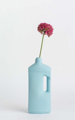 Prikazana je vaza brenda foekje fleur u svijetlo plavoj boji ispred sive pozadine. Vaza ima oblik kontejnera za sredstvo za pranje suđa. U vazu je utaknut pink cvjet.