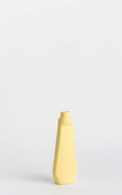 Prikazana je vaza brenda foekje fleur u svijetlo žutoj boji ispred sive pozadine. Vaza ima oblik kontejnera za sredstvo za pranje suđa. Vaza je prikazana lagano zaokrenuto na bočnu stranu.