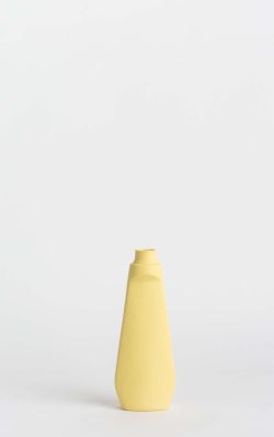 Prikazana je vaza brenda foekje fleur u svijetlo žutoj boji ispred sive pozadine. Vaza ima oblik kontejnera za sredstvo za pranje suđa.
