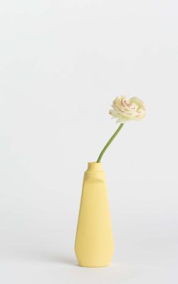 Prikazana je vaza brenda foekje fleur u svijetlo žutoj boji ispred sive pozadine. Vaza ima oblik kontejnera za sredstvo za pranje suđa. U vazu je utaknut bijeli cvjet.