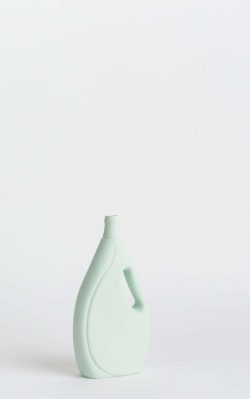 Prikazana je vaza brenda foekje fleur u svijetlo zelenoj boji ispred sive pozadine. Vaza ima oblik kontejnera za sredstvo za pranje suđa. Vaza je prikazana lagano zaokrenuto na bočnu stranu.
