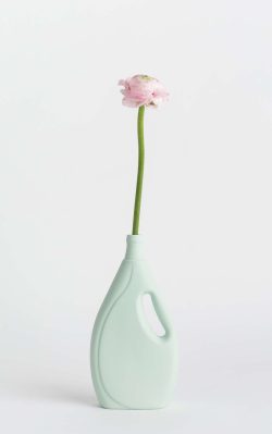 Prikazana je vaza brenda foekje fleur u svijetlo zelenoj boji ispred sive pozadine. Vaza ima oblik kontejnera za sredstvo za pranje suđa. U vazu je utaknut ružićasti cvjet.