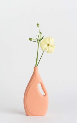 Prikazana je vaza brenda foekje fleur u svijetlo narančastoj boji ispred sive pozadine. Vaza ima oblik kontejnera za sredstvo za pranje suđa. U vazu je utaknut žuti cvjet.