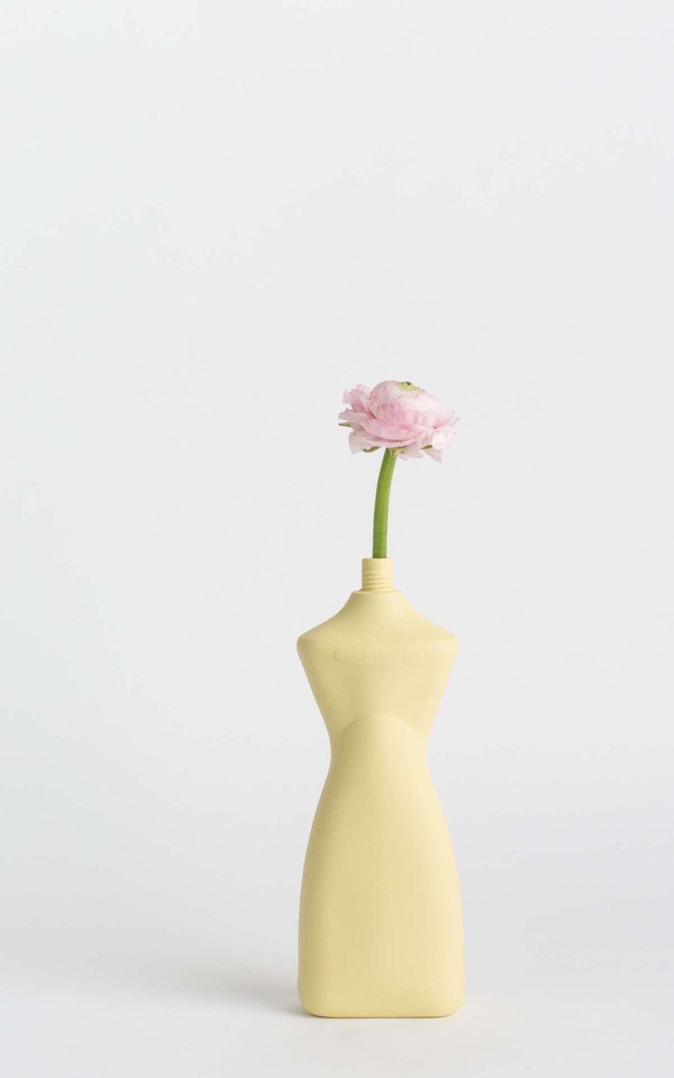Prikazana je vaza brenda foekje fleur u svijetlo žutoj boji ispred sive pozadine. Vaza ima oblik kontejnera za sredstvo za pranje suđa. U vazu je utaknut ružićasti cvjet.