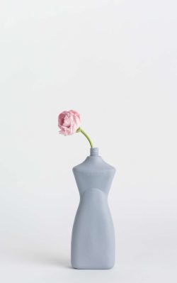 Prikazana je vaza brenda foekje fleur u svijetlo ljubičastoj boji ispred sive pozadine. Vaza ima oblik kontejnera za sredstvo za pranje suđa. U vazu je utaknut ružićasti cvjet.
