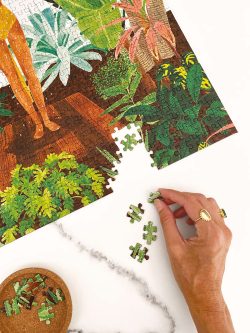 Ruka slaže puzle s motivom djevojke koja se tušira okružena biljaka na bijelom stolu
