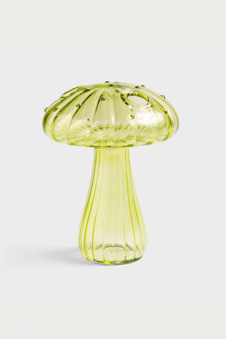 Na slici je staklena vaza u obliku gljive zelene boje. Vaza ima nekoliko rupica za cvjetiće na klobuku, stoji na sivoj pozadini