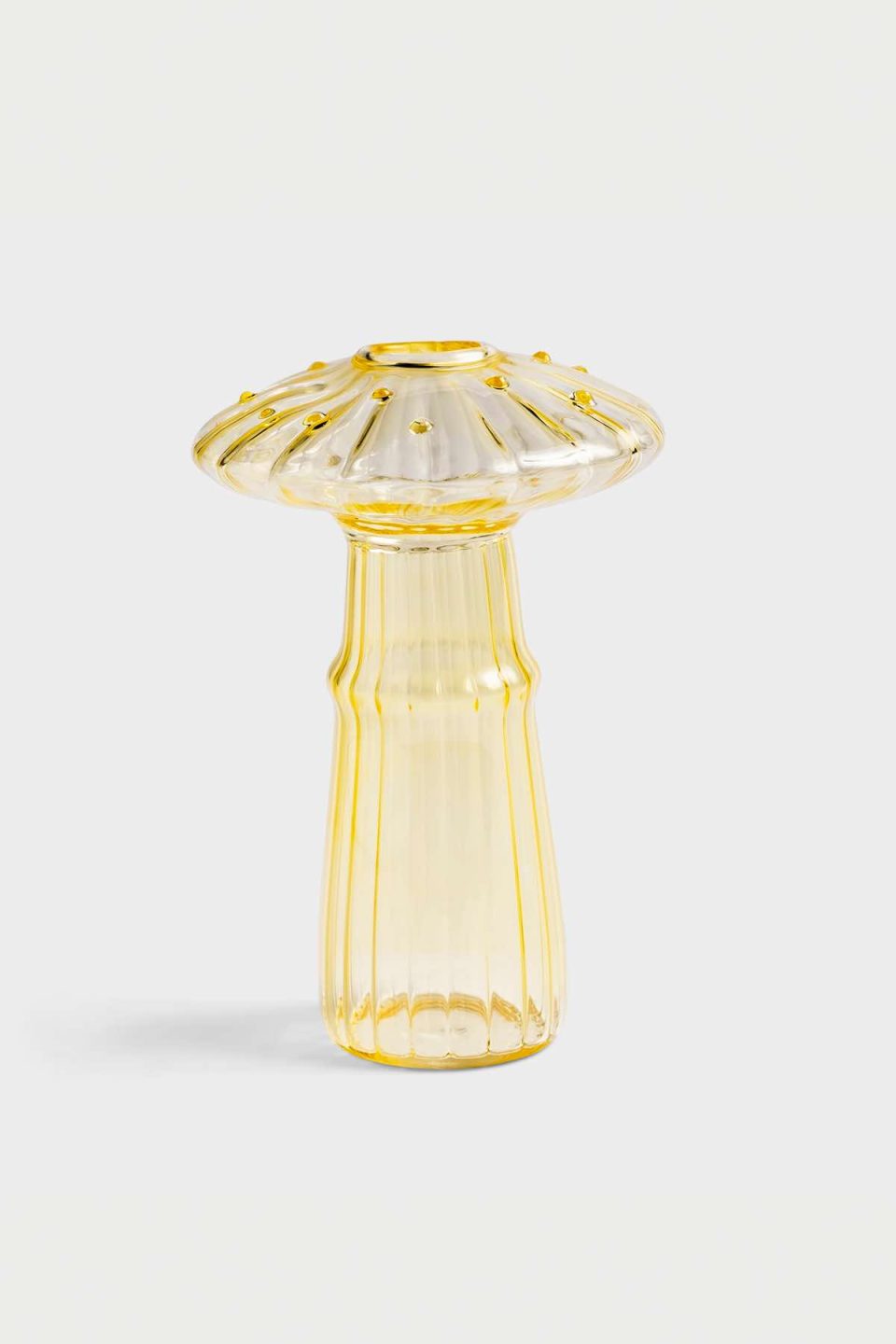 Na slici je staklena vaza u obliku gljive žute boje. Vaza ima rupicu za cvjetiće na vrhu klobuka, stoji na sivoj pozadini