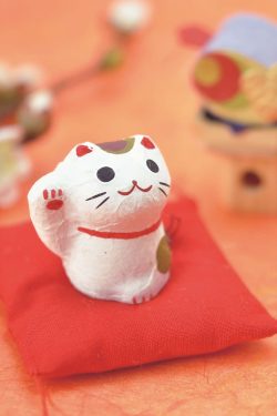 Na slici se vidi mala figurica mačkice od papirmašea u biljeloj boji na crvenom platnenom jastučiću. Mačkica drži desnu šapu u vis, u znak sreće. Ima crvenu ogrlicu i smješak na licu. Pozadina je narančaste boje, vide se nejasni obrisi grane sa cvjetovima trešnje i japanske dekoracije.