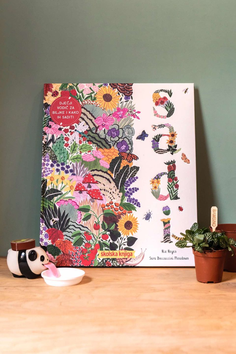 Na slici se vidi knjiga "Sadi". Naslovnica je bogato izilustrirana raznim biljkama i cvijećem. Knjiga stoji na drvenom stolu, naslonjena je na sivu pozadinu. Uz knjigu s desne strane je teglica sa sobnom biljkom, a se lijeve strane se vidi peropon u obliku pande.