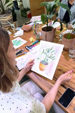 Slika prikazuje scenu s radionice crtanja. Ženska osoba drži list papira sa svojim crtežom u ruci. Crtež je izrađen s vodenim bojicama i prikazuje biljku. Osoba sjedi za drvenim stolom na kojemu se vidi pribor za crtanje, grickalice, pića i razne sobne biljke.