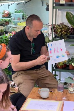 Slika prikazuje voditelja radionica crtanja biljaka kako objašnjava miješanje vodenih boja na listu papira koji drži u ruci i pokazuje polaznicima. Na stolu se vidi pribor za crtanje i pića.