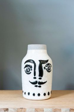 Slika prikazuje vazu od keramike ispred sive podloge na drvenom stolu. Vaza je glazirana bijela, ima na sebi nacrtano lice s brkovima u crnoj boji.