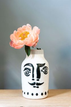 Slika prikazuje vazu od keramike ispred sive podloge na drvenom stolu. Vaza je glazirana bijela, ima na sebi nacrtano lice s brkovima u crnoj boji. U vazu je utaknut božur u blijedo rozoj boji.
