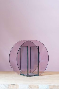 Na slici se vidi staklena okrugla vaza u rozo-ljubičastoj boji. Vaza stoji ispred roze pozadine na drvenom stolu.
