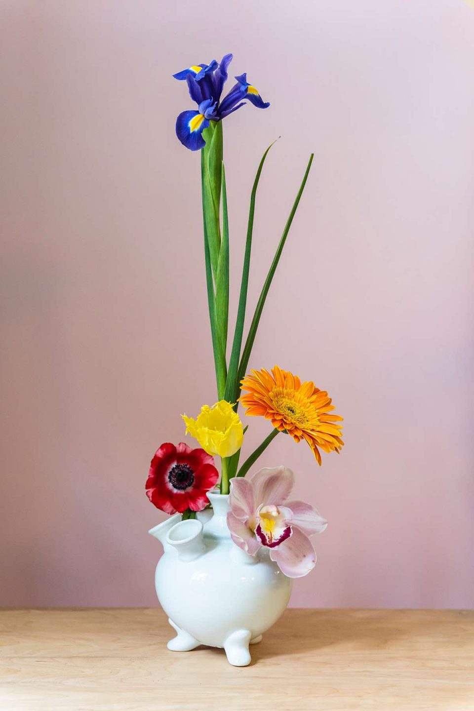 Na slici je prikazana vaza od bijelog porculana. Vaza je trbušasta s nekoliko otvora za cvijeće na vrhu. U otvore su umetnuti razni pojedinačni cvjetovi - gerberi, tulipan, orhideja i jedan jako visoki ljijan. Vaza stoji ispred roze pozadine na drvenom stolu.