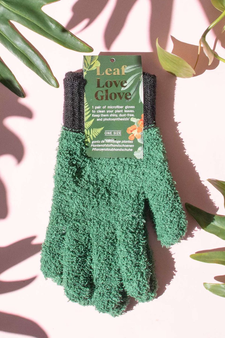 Na slici se vidi par zelenih rukavica od frote materijala na rozoj pozadini okružene listovima biljaka. Na etiketi piše "Leaf Love Glove", što upućuje na to da se radi o rukavicama za brisanje prašine s listova.