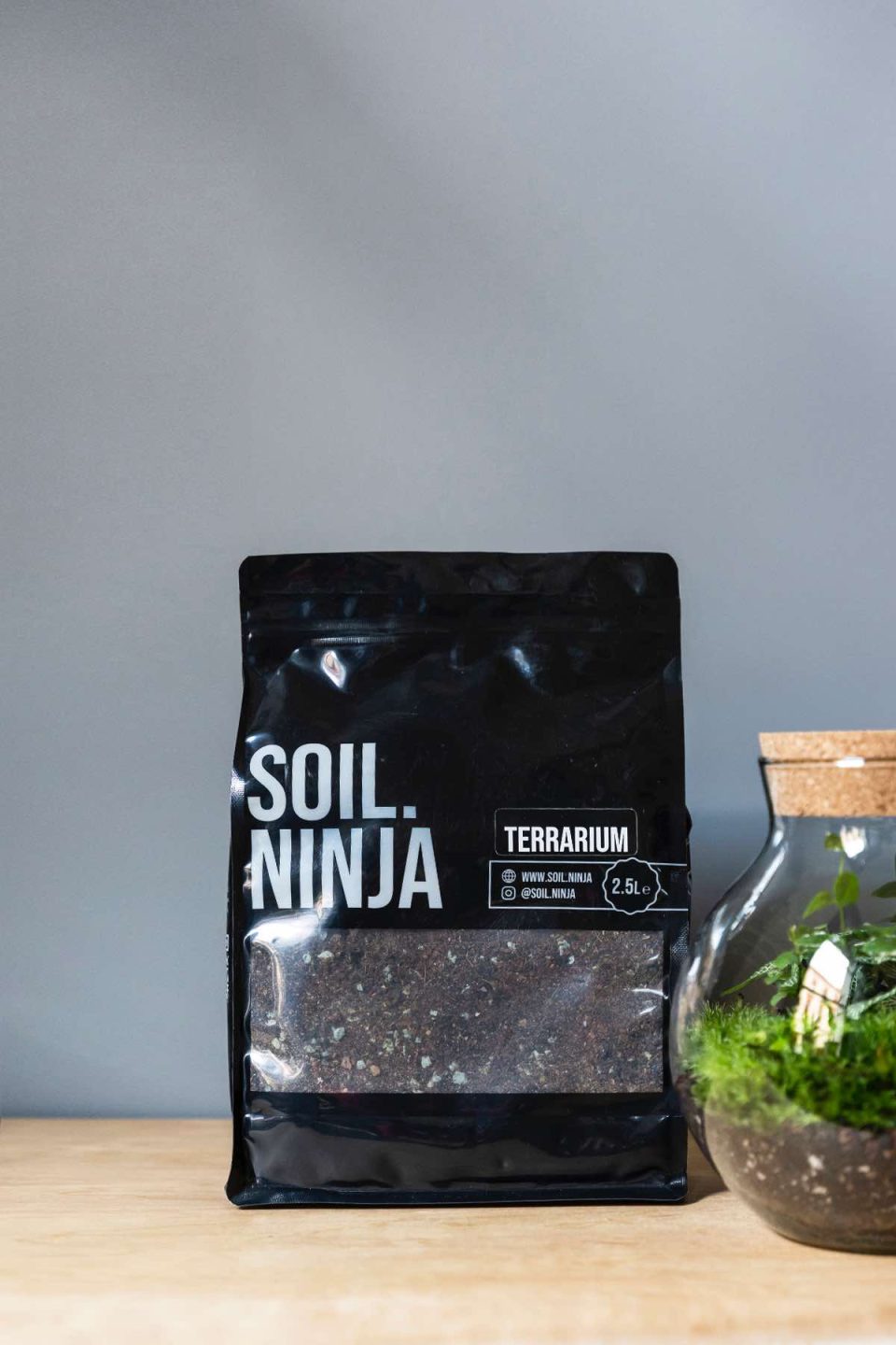 Na slici se vidi vreća supstrata od 2,5 kg proizvođača Soil.Ninja za terarihe. Uz vreću stoji biljni terarij u okrugloj staklenci.